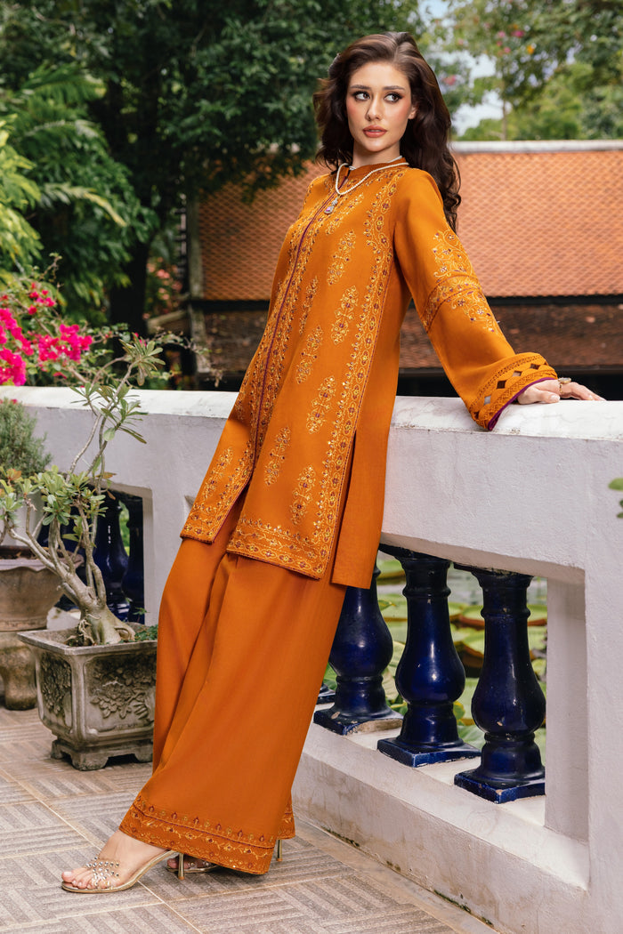 Woman in an orange traditional outfit standing outdoors with greenery and a building in the background.