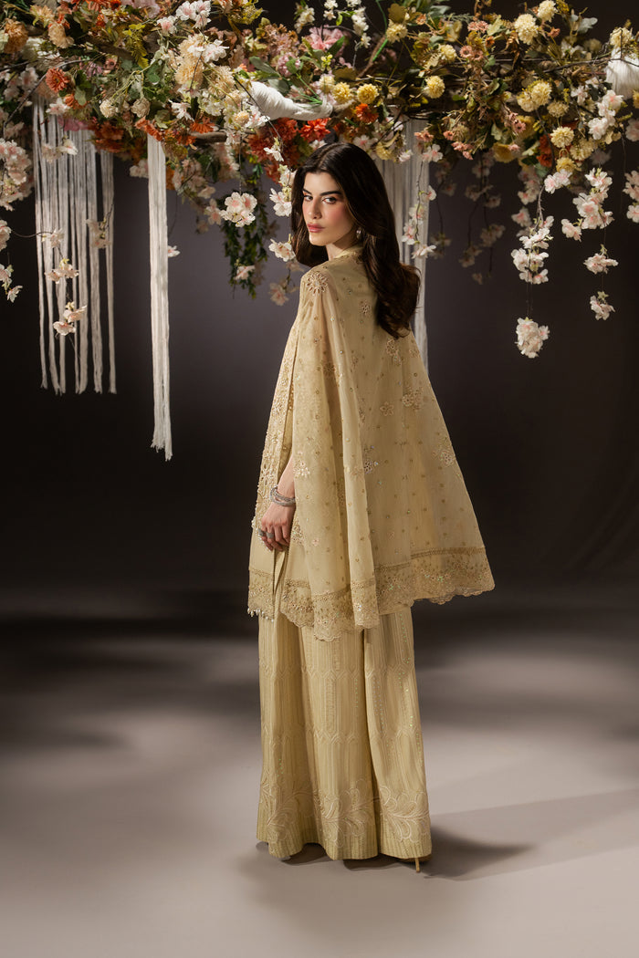 Three-quarter back view of Livia highlighting the draped pale yellow Organza cape and full-length wide flare pants.
