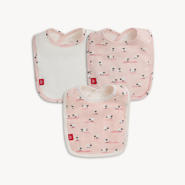 A three-pack of baby bibs featuring one solid white bib with a light pink border and two bibs with a repeating print of white sheep on a light pink background, By Magnetic Me.