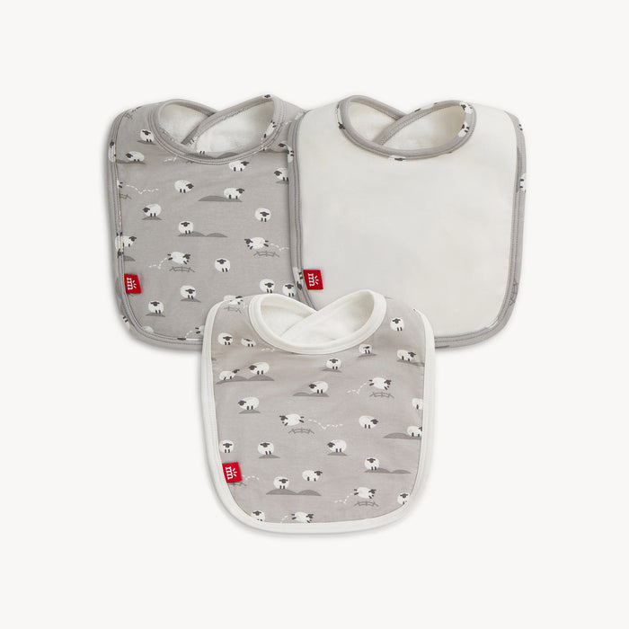 A three-pack of baby bibs featuring one solid white bib with a light gray border and two bibs with a repeating print of white sheep on a light gray background, By Magnetic Me.