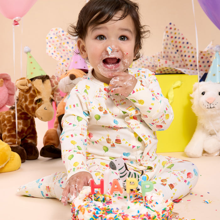 Child celebrating a birthday with cake and balloons in a colorful room wearing A cream-colored long-sleeved convertible coverall with a watercolor "It's My Birthday" print and hidden magnetic closures that easily swap between footed and footless styles, by Magnetic Me.