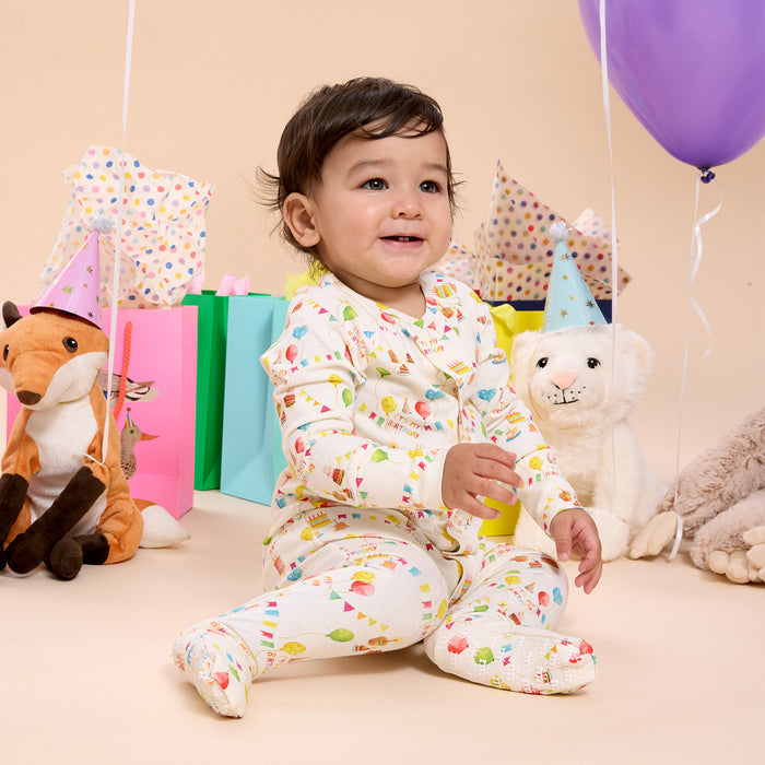 Child surrounded by party decorations and stuffed animals on a beige background wearing A cream-colored long-sleeved convertible coverall with a watercolor "It's My Birthday" print and hidden magnetic closures that easily swap between footed and footless styles, by Magnetic Me.