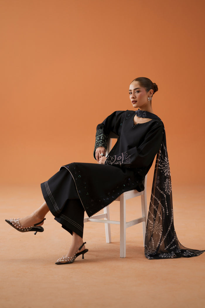 Seated model wearing Riedel black embroidered ready-to-wear outfit with printed chiffon dupatta and culotte trouser for semi-formal styling.